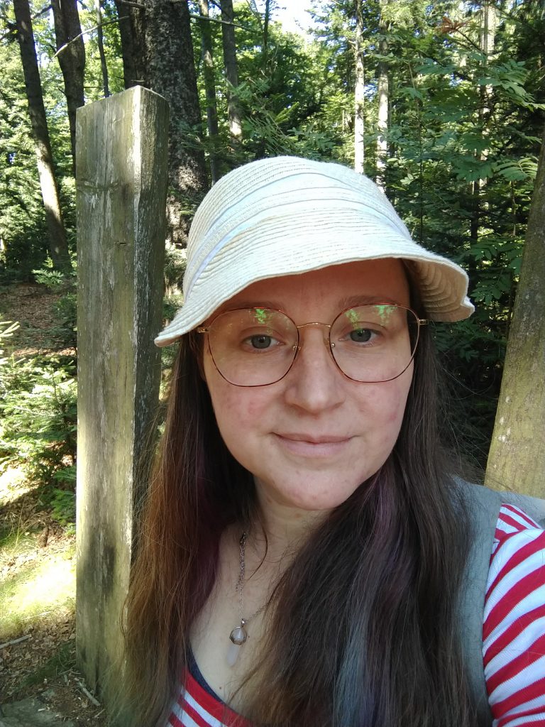 Eva steht im Wald, außenrum Bäume. Sie trägt ein rot-weiß gestreiftes Oberteil, eine geflochtene Kappe und eine Brille. Sie lächelt in die Kamera.